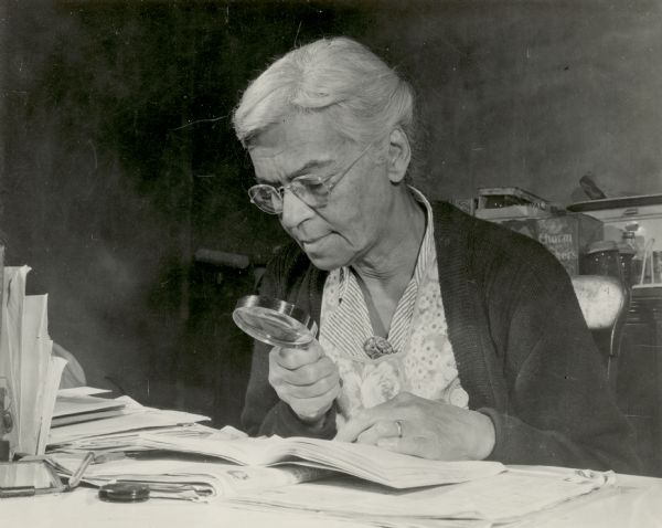 Portrait of Dr. Lillie Rosa Minoka Hill, a physician of Mohawk ancestry. She is sitting at a table examining a book through a magnifying glass. Dr. Minoka Hill married Charles Hill, an Oneida man, and moved with him to the reservation in Wisconsin. She was adopted by the Oneida in 1947 and became a prominent doctor in the state.