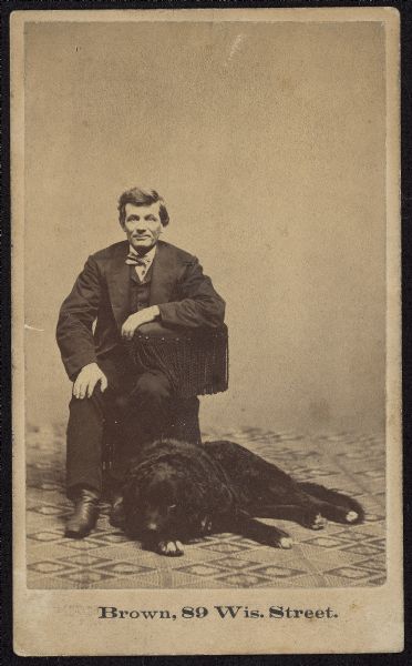 Carte-de-visite of Joseph Mabbett Allcott, proprietor of J.M. Allcott & Co. drug store in Milwaukee. He is sitting in a chair, with a dog resting at his feet. Caption on back reads: "Proprietor of first true drug store (in modern sense), Milwaukee."