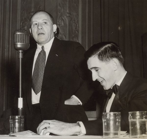 View of a man standing at a microphone. A man is seated at a table next to him and appears to be laughing. Caption reads: "Col. Robert S. Allen, Washington newspaperman, former Merry-go-Round columnist, speaking in April 1947. Former reporter for Capitol Times."
