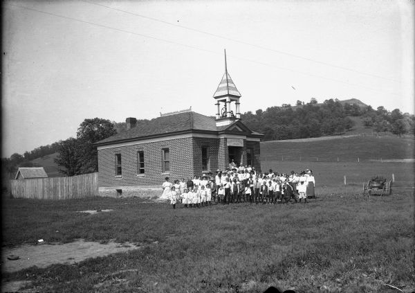 View of a large group of people standing in front of a brick building with a bell tower. The sign above the entrance reads: "Public School 1905." A fence runs from the rear of the building on the left, and a cart is on the far right. Behind the school is a hill and trees. Caption reads: "Lone Rock, Wis. (?) c. 1905-1910. Public school and pupils."