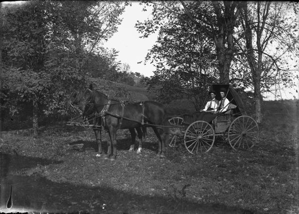 View of two people seated in a buggy. Two horses are hitched to the buggy. A steep hill is in the background on the left.