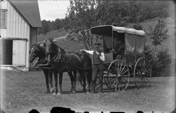 View of a man standing and posing with a horse-drawn covered wagon. Another man is sitting in the wagon. A building is in the background on the left. On the right is a steep hill and trees.