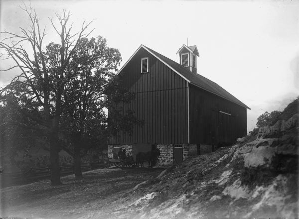 View towards a barn with a man and another person sitting in a horse-drawn wagon near the slightly open barn door. A split rail fence runs along the left to the back of the barn, and in the background are corn shocks in a field, and beyond is a steep hill. In the right foreground is the rocky base of a hill.