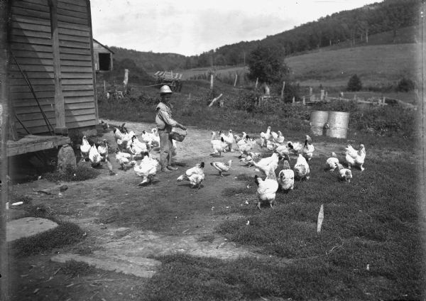 View of a boy standing barefoot outside a farm building holding a container. A flock of chickens surrounds him. In the background is a fence, fields, and a hill.