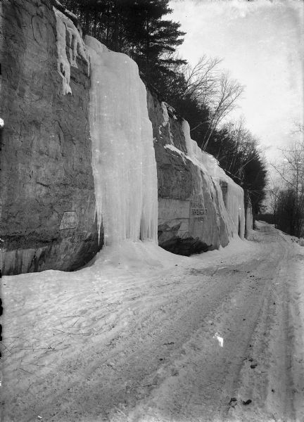 View down snowy road along a bluff with frozen ice falls on the rock faces. Two signs are painted on the bluff. One reads: "Painting and Paper Hanging, R. Culver." The other reads: "Reliable and Responsible Implements, J.F. Beardsley."