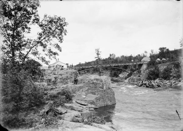 Elevated view from left side of a rocky shoreline of the Wisconsin River toward a bridge spanning it. Two men are standing on the rocks in front of the bridge just above a boat resting on the rocks below. Logs are piled in the water near the base of the bridge on the right. Buildings are on either side of the bridge, and two men are standing on the bridge outside the building on the left.