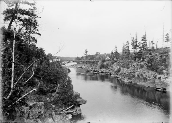 View of the Wisconsin River, with a bridge in the distance. Buildings are on both sides of the bridge, and logs are piled below the bridge on the right.