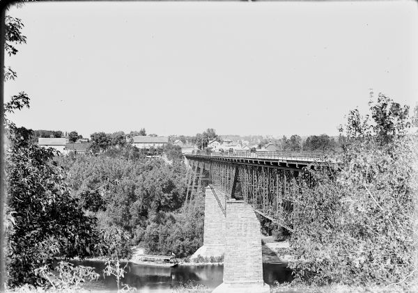 View across the Wisconsin River towards buildings on the other side, with a bridge on the right supported by brick-covered supports. An excursion boat is far below along the opposite bank. Pedestrians and horse-drawn vehicles are on the bridge.