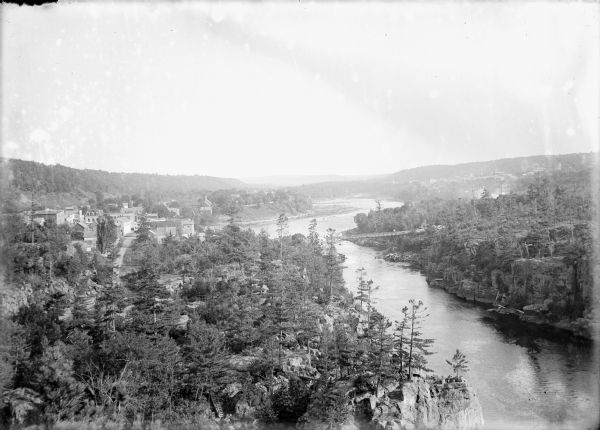 Elevated view of the town of Lone Rock on the left, and the Wisconsin River nearby on the right. In the foreground below is a small bridge leading over a ravine toward a bench on a rocky overlook of the river. A bridge spanning the river is near the town. Logs are at the base of the bridge on the right riverbank, and small buildings are at each end of the bridge. Another town is across the river in the background on the right.
