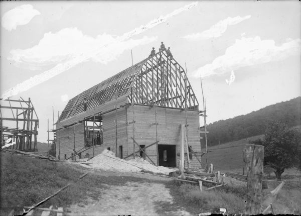 View of a barn under construction. The roof support beams are in place. Several men are posing on the left in the hay loft and one man above standing on top of the wall. Three men are posing on the roof supports at the front of the barn. On the right is an open field on a hill, with trees in the background. On the left is a smaller building that also appears to be under construction.
