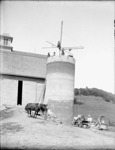 View of a silo. A man with a horse is standing at the base of the silo on the left, and a man is standing near a wagon on the right. At the top of the silo several men are posing with rigging at the top. The top of the silo not yet been domed. A barn is behind them on the left, and a field and a hill are in the background on the right.