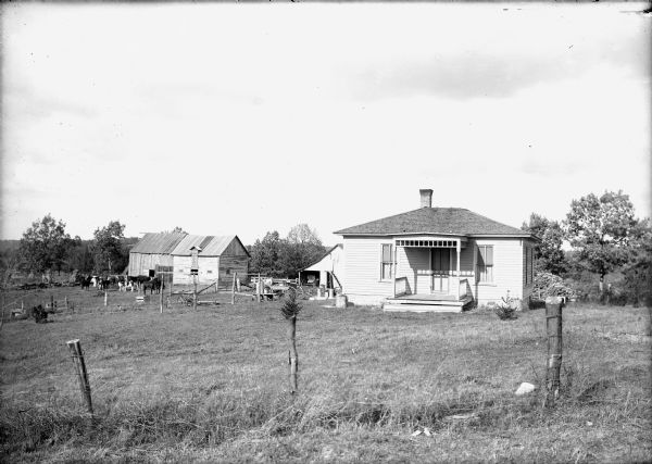 View over fence towards a farmhouse and barns, as well as yards enclosed by wire fencing. Animals are outside the barn. A woman is standing between the building behind the house on the left, and the barns on the far left.