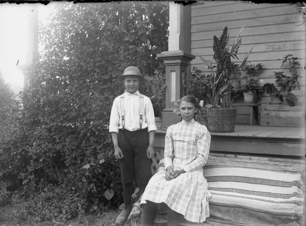View of a boy and a girl posing in front of a porch. The girl is sitting on the steps which are covered with a cloth, and the boy is standing on the left. There are potted plants on the porch behind them.