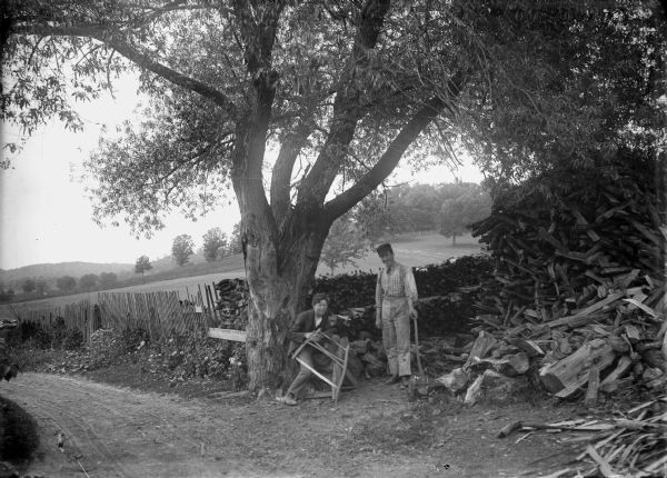 Two young men are posing under a tree in front of neatly piled stacks of fuelwood, and a tall pile of fuelwood on the right. The man on the left is sitting and holding a bucksaw, and the man in the center is standing and holding an ax. The road in the foreground leads to the left and runs along a fence and fields. In the distance is a tree-covered hill.
