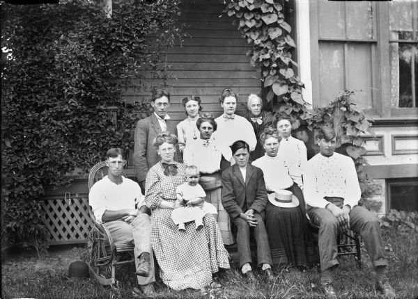 View of twelve people, men, women, and children, posing together in front of a building.