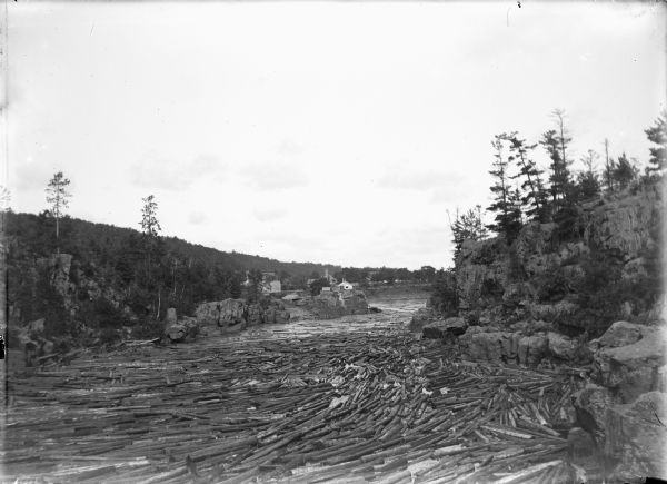 Elevated view of logs floating down the Wisconsin River, with rocky cliffs along the shoreline. A bridge and buildings are in the background, with a tree-covered hill along the left.
