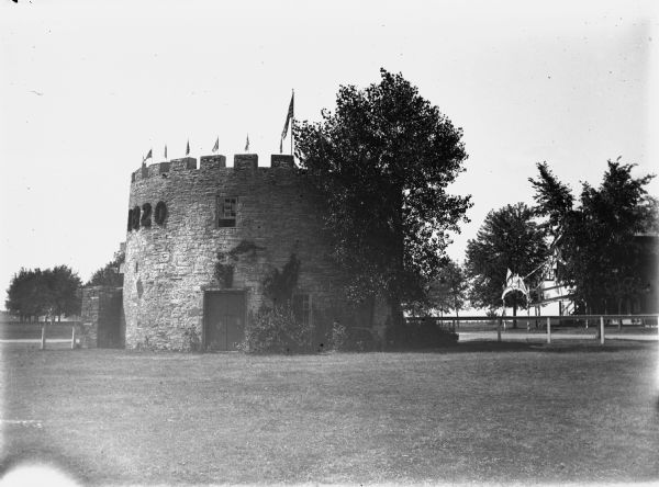 View of a post tower at Fort Snelling. A tree partially obscures the tower. Flags are arrayed on the battlements and the number "1820" (the year fort construction began) is on the left side of the tower. There is a building in the background on the right, and an arch with decorations is over the front walk to the building. Caption reads: "Minneapolis, Minn (?). c1905-1910. Fort reconstruction?"