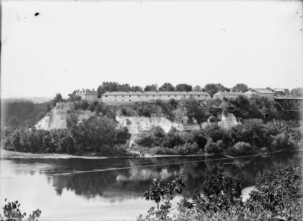 Elevated view across river from the top of a cliff towards buildings along the the top of the cliffs on the opposite side. Far below a group of people are standing on the sandy shoreline. There is a bridge on the far right, and just beyond it is a building or arch decorated with the words: "Welcome D.A.R." The cliffs below the buildings have been reinforced with stone. The river bends around to the left, and a long, wooden staircase on the left leads from the buildings down to treeline at the base of the cliff. Caption reads: "Lone Rock (?), Wis. c1905-1910. View of Wisconsin River and buildings on the bluffs."