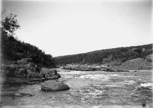 Slightly elevated view of a river with choppy waves. The shoreline is rocky on the left and a man is sitting on one of the rocks. Further down the river on the right is a group of people sitting on part of a pile of large logs stacked on the shoreline. The hills on both sides are tree-covered, and buildings obscured by trees are partially uphill on the far right. Caption reads: "Lone Rock(?), vicinity, Wis. c1905-1910. Log jam(?) on the Wisconsin River."