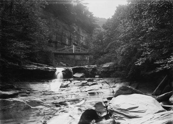 View upriver across rocks towards water falling over a rock under a small wooden bridge. The rockface of a bluff rises above the bridge in the background, with another tree-covered bluff behind it. Caption reads: "Lone Rock(?), vicinity, Wis. c1905-1910. Falls in the Wisconsin River(?)."