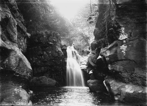 View toward a waterfall from the river below. Rock faces rise on both sides, and trees are growing along the tops of the cliffs. Caption reads: "Lone Rock(?), vicinity, Wis. c1905-1910. Falls in the Wisconsin River(?)."