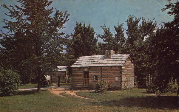 View across lawn towards two log structures among trees, and an ox-drawn wagon on the left side of the building in the front. Postcard text reads: "<b>DR. JOHN ALLEN'S RESIDENCE</b> Doctor John Allen came to New Salem in 1830. In 1833 he built a three room log residence and on March 27, 1834, married Mary E. Moore. Because of his extensive practice he became one of New Salem's most prosperous citizens. He was a devout Presbyterian and established the village's first Sunday School and Temperance Society in his house.
The Allens moved to Petersburg in 1838. The following year a tenant whose name now unknown lived in the cabin; the next two years the Traylor family occupied it and finally, in 1842, it was abandoned.
The main room was the parlor, dining room and kitchen, the leanto served as a bedroom and the west room was used by the doctor as an office.
Inn the background is the ox team and Conestoga wagon, and Rutledge Tavern."