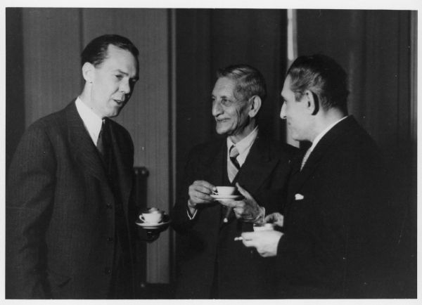 Three men standing together holding cups and saucers. Two have cigars or cigarettes. Caption reads: "George V. Allen (left) with the Foreign Minister of Iran and the Chief of Protocol (1948)."