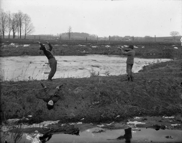 Staged scene of a man pointing a rifle at another man, who has thrown his hands up and is leaning back. A third person is lying on the ground. A pond is behind them, and water is in the foreground. Caption reads: "Mayville, Wi. Comic shooting scene; man shoots man with rifle."