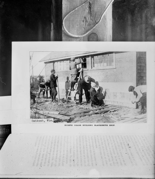 A book is opened to a photograph of children working around the exterior of a building, while two men are looking on. Photograph is captioned: "Eighth grade building blacksmith shop."