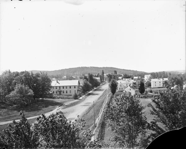 Elevated view of a town, looking down from the right side of the main street and several buildings. In the distance are tree-covered hills. Caption reads: "Scandinavia (?), Wis. c.1915-1930. View of town."