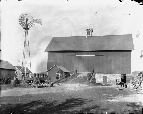 Side view of a barn, with a ramp leading up to its door. A windmill is on the left just behind a wagon loaded with bins or barrels. Other farm buildings are in the background on the left. The sign on the windmill reads: "The Aermotor Chicago."