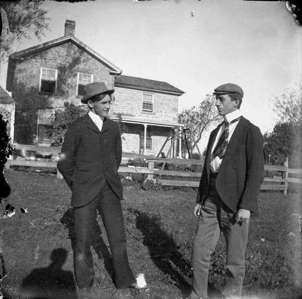 Two young men are standing in the foreground. Behind them is a fence and a house. There is a yard swing in the yard on the right. Caption reads: "Madison, Wis. c.1910-1915. "Clarence [Scott Hean] and Robbie G." Findlay house. Corner Ridge and Harvey, approx. 3000 block University Ave."