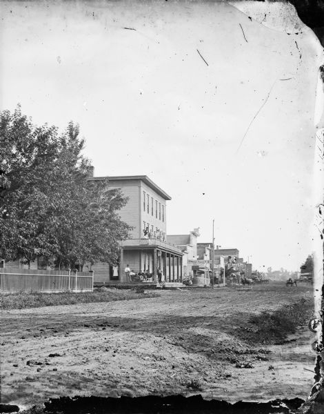 View across unpaved road towards a row of buildings on the other side. A fence and trees are on the left, and in front of the first building a group of men are sitting on the porch, two children are sitting on the steps, and a group of men and women are on the balcony above. Other buildings have signs that read "Howard and Hansen Hardware," "European Restaurant" and "Drug Store." Horse-drawn vehicles are along the curb and in the road. Caption reads: "River Falls, Wis. About 1890(?). View of a main street, with inn(?) in foreground."