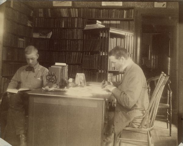 Professor William F. Allen seated and writing at a desk in the study of his home at 228 Langdon St. His son Will is seated in a chair and reading. A large book shelf can be seen against the wall.