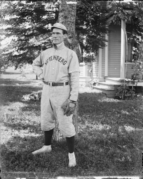 A man is standing and posing in a Wittenberg Grays baseball uniform and wearing a catcher's mitt. He is standing in a yard under a tree, and a building with a porch is in the background.