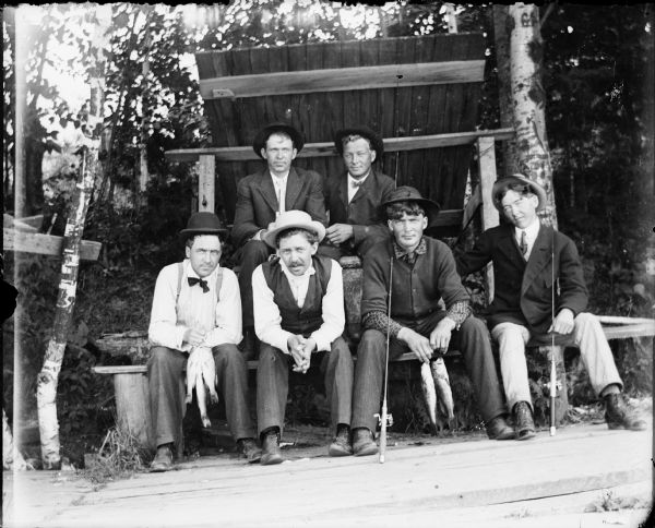 Six men posing sitting in front of a lean-to. They are holding fishing poles and fish.
