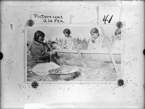 Three women are kneeling and binding birch bark to a canoe frame. A girl is standing behind the canoe in the center. Buildings are in the background.
