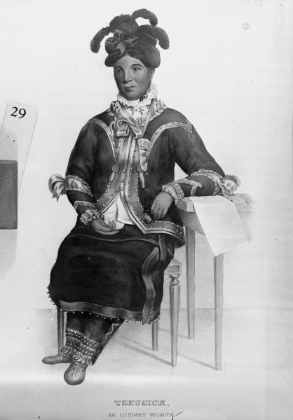 Portrait of a woman, who is sitting with her left arm resting on a desk or table. She is wearing a jacket, hat, skirt, and pants. According to <i>History of the Indian Tribes of North America</i> by Thomas L. McKenney & James Hall, Tshusick was an Ojibwa convert to Christianity who impressed many members of Washington, D.C. society in the 1820s. She claimed to have walked there from Detroit. However, the authors learned that she was a wanderer and prone to creating fanciful stories in her adventures.