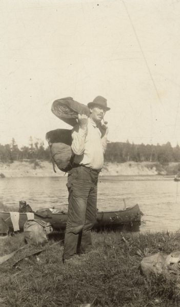 UW-Madison sociology professor Edward Alsworth Ross posing on a river bank. He is wearing a hat and backpack, and is holding a bedroll and has a pipe in his mouth. A canoe is on the shore behind him. Caption reads: "Edward Alsworth Ross. Canoe trip in Ontario, Canada, circa 1920. (reproduced opposite page 250 in Ross' autobiography, "Seventy Years Of It." published D. Appleton-Century Company, New York, 1936)."