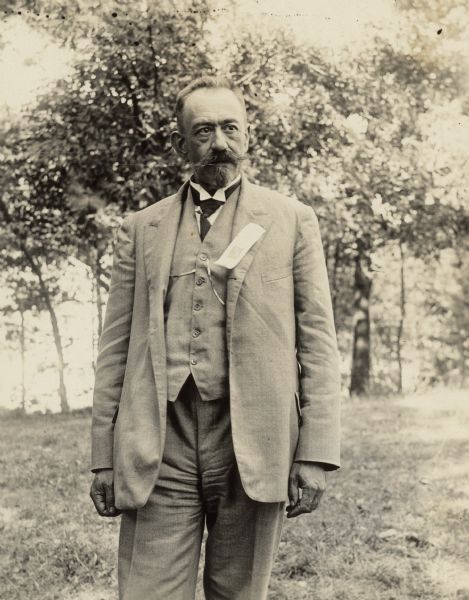 Three-quarter length portrait of Alois Alphonsus, of the Austrian Department of Agriculture, standing outdoors. Alphonsus was a beekeeper who came to Madison in the 1920s. He is wearing a suit with a ribbon or tag attached to his lapel.