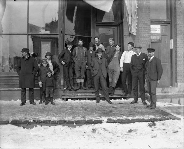A group of people pose outside a building. Signs in a doorway advertise the North British and Mercantile Insurance Company, and the Hartford Fire Insurance Company.