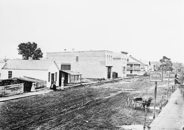 Elevated view of an unpaved main street, with the Village House across the street in the center background. Wooden buildings and wooden sidewalks are on both sides of the street. Across the street on the left two women are standing on the sidewalk, and another woman is standing near a shed which appears to be full of fuelwood. In the foreground a horse and wagon are parked next to a sign for the DePere House on a post above the sidewalk. Further down on the right is a sign shaped like a shoe, and further down is a sign shaped like a shirt or coat. Caption reads: "South Broadway about 1870 (business section) DePere. Village House oldest hotel in this section kept by W. P. Call."