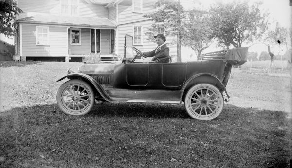 A man poses in the driver's seat of a car in the yard in front of a farmhouse with a porch. Caption reads: "Man in open touring car in front of farmhouse."