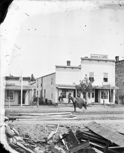 View across an unpaved street of a building with three storefronts. Signs on the taller building on the right read "Drugstore," "Books/Stationery," and "Photograph Gallery." Two men holding their hats are standing outside the storefront on the left side of the building under the porch roof, which has a support pole and a post painted like a barber's pole. Across the alley on the far left is a building which has a large round clock on a pole suspended over the wooden sidewalk. In the street in the foreground is a woman who is riding sidesaddle on a horse. Caption reads: "River Falls, WI. Drugstore, stationery store, photo studio, all in one frame building on a main street. East Main just south of Maple."