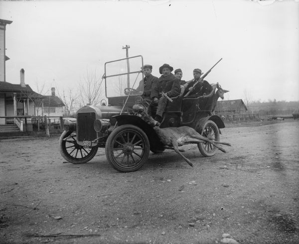 Four men posing in a car on a road, with a man sitting behind the steering wheel on the right side. The car has a license plate on front that reads: "5083 Mich." The two men on the left side of the car facing the camera are holding rifles. A deer is fixed to the running board, and a second deer is to be fixed to the rear of the car on the right. In the background are buildings.