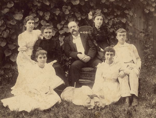 Group portrait of two men and five women. On the ground in the center are hand fans, and a hat. Caption reads: "The Simons Family. Left to right, seated on chairs: Sarah E. Simons, Emma Comstock Simons, Mr. [Frank] Simons, Mary Simons, Frank Simons. Left to right, seated on ground: Daisy Simons, Rosamond Simons. Sarah E. Simons never married. Mary Simons married Ernest Lent. Daisy Simmons married Victor Mason. Rosamond Simons married Edward A. Ross."