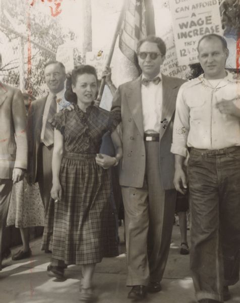 Several people marching together. A man and woman are marching in the front next to each other; the woman is holding a cigarette. A sign reads: "[Danville] can afford a wage increase. They [?] 12 1/4 [?] last [?]." Caption reads: "A WIFE'S PLACE is at her husband's side, especially when the going gets rough, so Mrs. George Baldanzi [Lena Parenti] walks the picket-line at Danville with TWUA's executive vice-president."