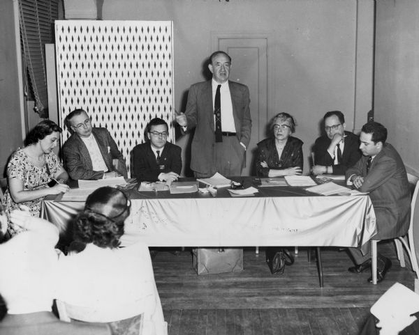 Five men and two women are at a table in front of an audience. One man is standing and gesturing. Caption reads: "DAY LONG SESSION on community services in the big town draws a delegation from Greater New York Joint Board (above) [not pictured here]. Below: Solomon Barkin, TWUA [Textile Workers Union of America] research director, takes the floor during discussion of the needs of older workers."