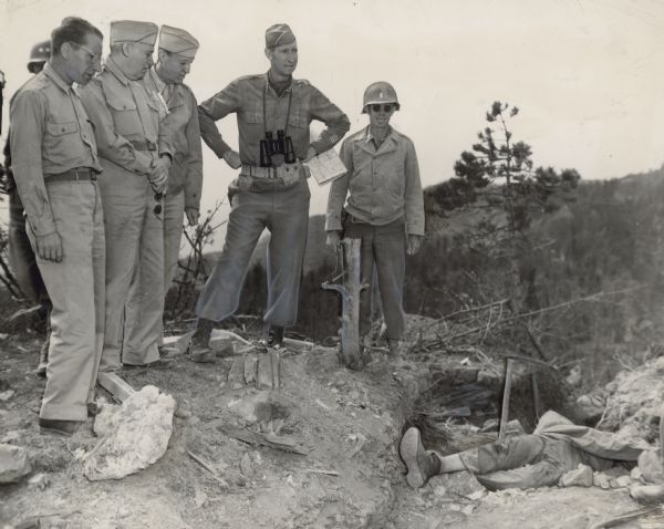 Five men in uniform stand over a partially obscured body on a hillside. Another soldier is behind the group on the left. Caption reads: "Fifth Army, Bonzalla Area, Italy. Lt. Gen. Mark W. Clark, C. G. Fifth Army, and Mr. Luigi Antonini, AFL, and Mr. George Baldanzi, CIO, U.S. Labor Representatives, look at German soldier killed in his dugout by artillery fire. Photo by Thomas - 3131 Sig. Sv. Co."