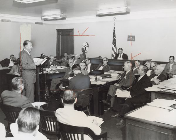 A man stands at a podium and reads from a sheet of paper. Several men are seated at tables and desks, and are looking at him. Caption reads: "PLEA FOR PUBLIC POWER in New England to stimulate industry is being made here by Solomon Barkin, TWUA research director, as CIO spokesman before a Hoover Commission task force on water resources. Barkin blamed private-utility lobby's greed for region's industrial decline. At desk in rear is Admiral Ben Moreell, presiding officer at the hearing. Glum-faced listeners are members of the task force."